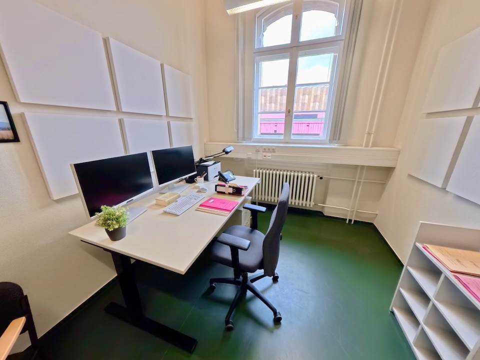 Ein modern eingerichtetes Büro in einem Altbau des Landgerichts Berlin II im Dienstgebäude Littenstraße. Zu sehen ist ein weißer, höhenverstellbarer Schreibtisch mit zwei Monitoren, Tastatur und Akten. An den Wänden hängen weiße Schallschutzpaneele.