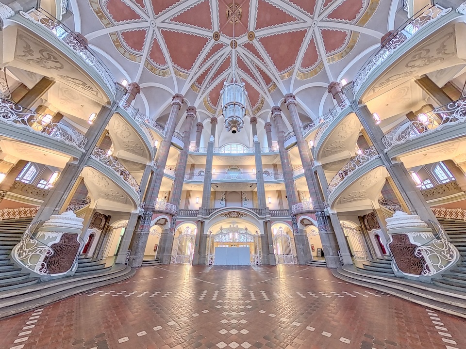 Imposante Eingangshalle des Landgerichts Berlin II im Dienstgebäude Littenstraße. Eine hohe Gewölbedecke mit rosafarbenen Feldern wölbt sich über umlaufende Galerien mit weißen Geländern und verzierten Säulen. Ein großer, zentraler Kronleuchter hängt herab.