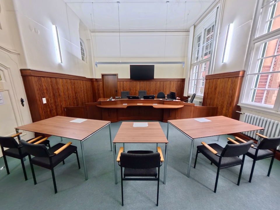 Blick in einen Gerichtssaal des Landgerichts Berlin II im Dienstgebäude Littenstraße. Im Zentrum stehen drei Tische in U-Form, davor dunkle Stühle. Im Hintergrund befindet sich der erhöhte Richtertisch auf einem Podest, umrahmt von dunkler Holzvertäfelung.