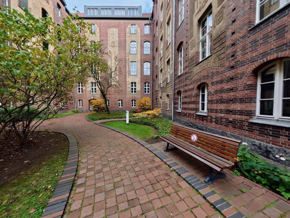 Ein Innenhof mit Rasenflächen, Bäumen und einem geschwungenen Pflasterweg des Landgerichts Berlin II im Dienstgebäude Littenstraße. Eine Holzbank mit einem Nichtraucher-Hinweisschild steht vor der detaillierten Backsteinfassade des Gebäudes.