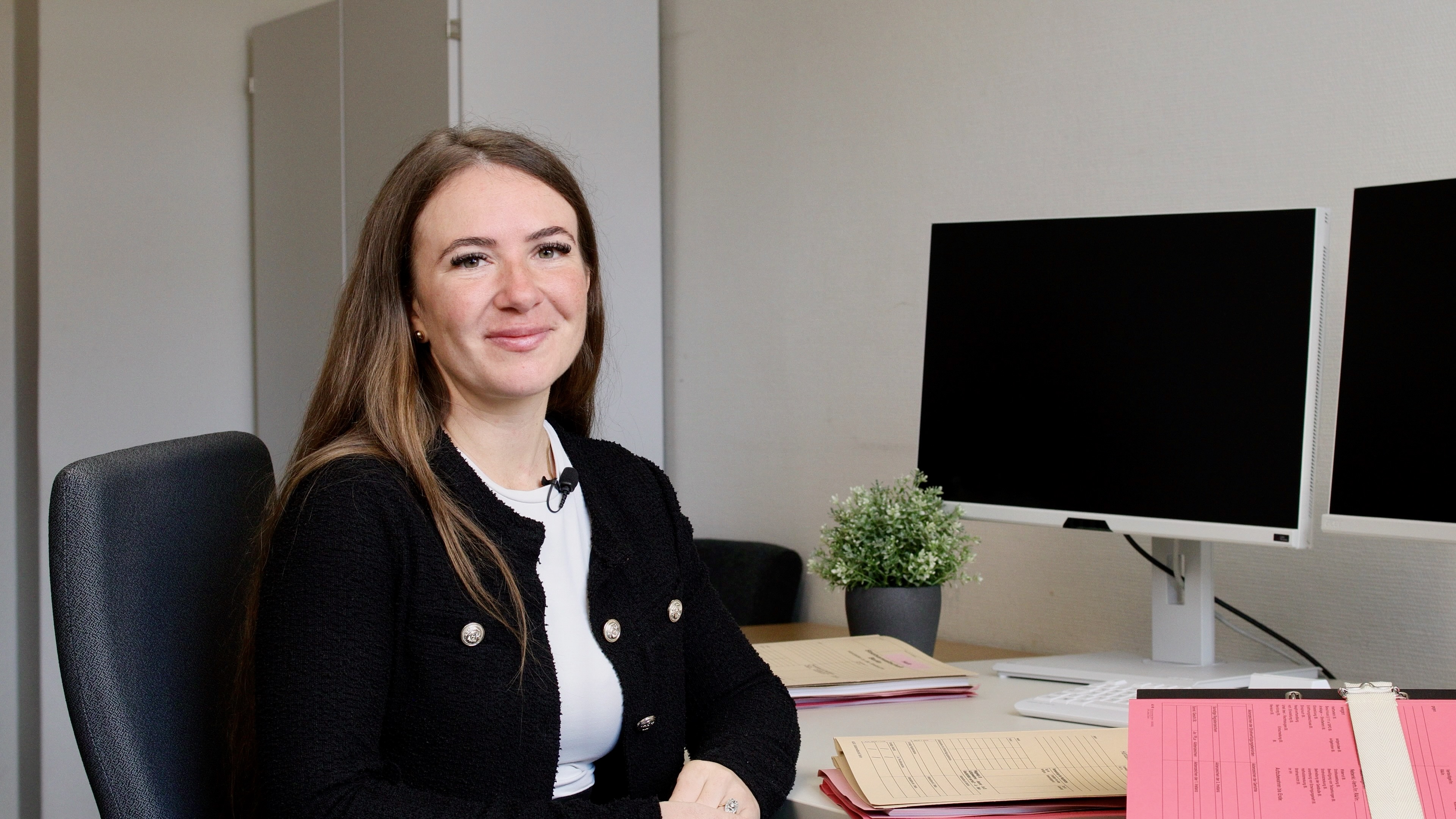 Porträt einer Justizfachwirtin am Schreibtisch. Sie lächelt in die Kamera, vor ihr liegen Arbeitsunterlagen und im Hintergrund sind zwei Computerbildschirme zu sehen.