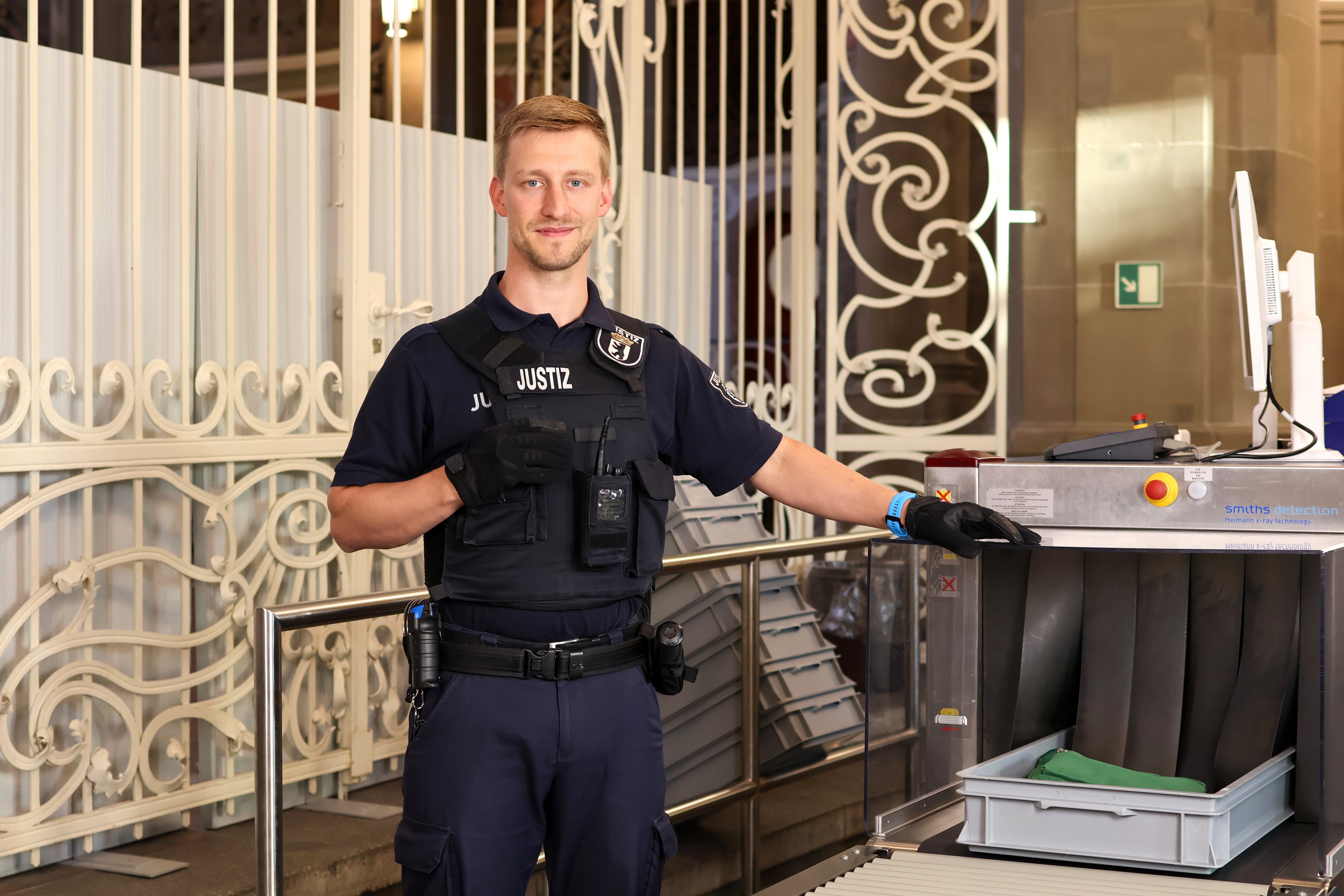 Ein Justizhauptwachtmeister in dunkelblauer Dienstkleidung steht neben einem Gepäckscanner im Eingangsbereich. Er trägt eine Schutzweste mit der Aufschrift 'JUSTIZ'.