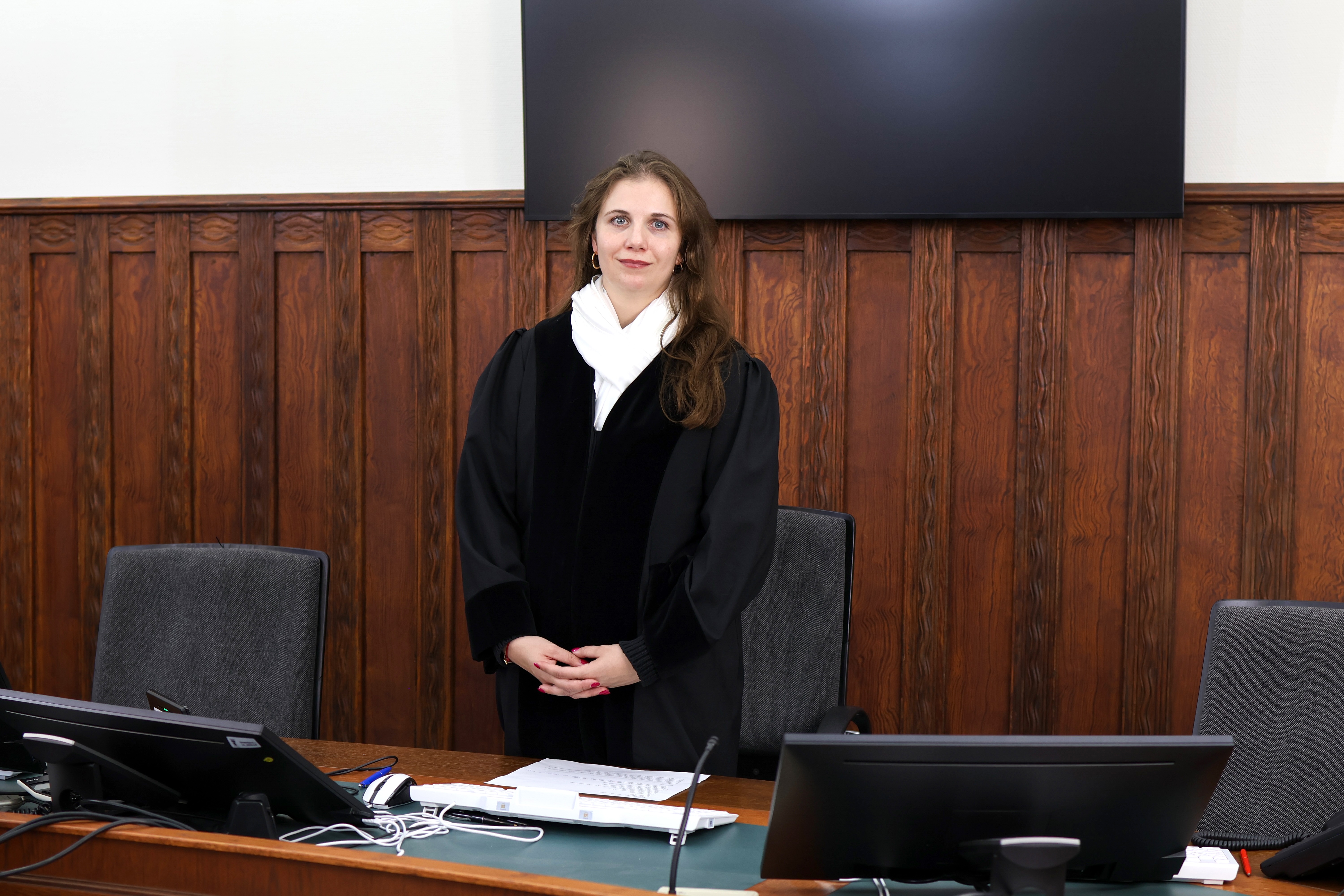 Eine Richterin steht in einem holzgetäfelten Gerichtssaal hinter dem Richtertisch. Sie trägt eine schwarze Robe mit weißem Halstuch und blickt in die Kamera.